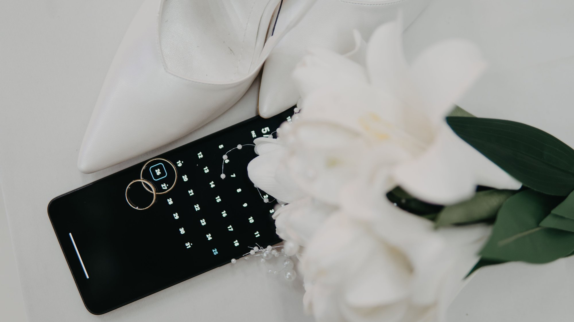 Celular com calendário aberto ao lado de sapato de noiva, alianças e buquê, simbolizando a escolha da data em alta temporada de casamentos